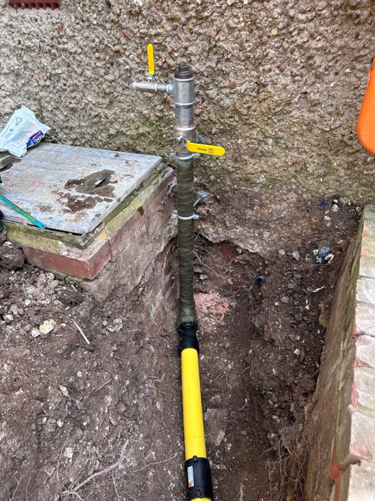Yellow gas riser with an isolation valve emerging from an excavation beside a flint wall